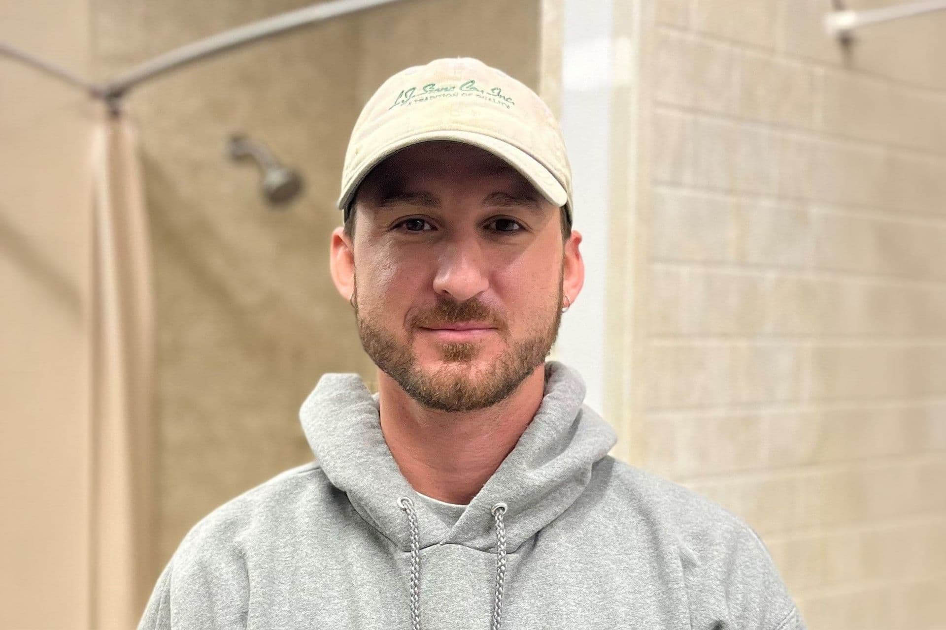 Man wearing cream-colored baseball cap and gray hoodie, smiling at camera in indoor setting
